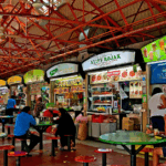 Screenshot 2025-11-24 153656 Suasana hawker center di Singapura dengan deretan meja makan, kios makanan sibuk, dan pengunjung yang menikmati hidangan lokal di area terbuka yang terang dan ramai