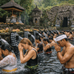 ritual melukat di bali Ritual melukat di Pura Goa Gajah Bali dengan peserta lokal dan wisatawan asing di bawah pancuran air suci, berlatar arca batu bersarung poleng dan pura kuno yang asri.